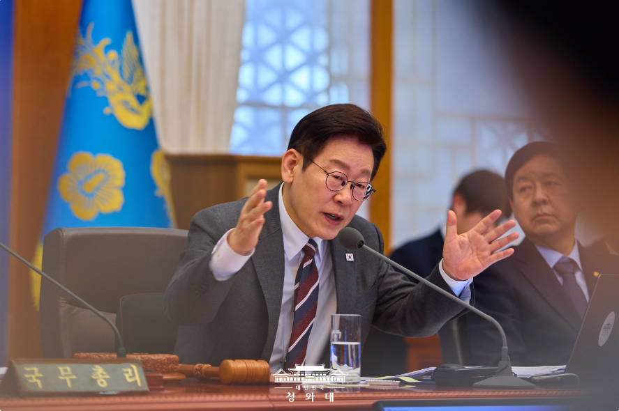 대한민국 청와대(또는 대통령실) 국무회의장에서 이재명 대통령이 안경을 쓰고 진지한 표정으로 양손을 사용해 발언하고 있는 모습. 배경에는 청와대 로고가 새겨진 깃발이 있으며, 앞에는 '국무총리' 명패와 의사봉이 놓여 있어 국가 핵심 정책을 결정하는 엄중한 분위기를 전달함.